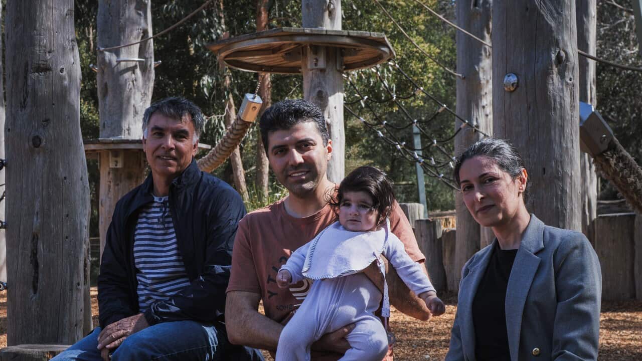 A woman and two men sit on a wooden structure at a playground. A man in the middle is holding a baby.