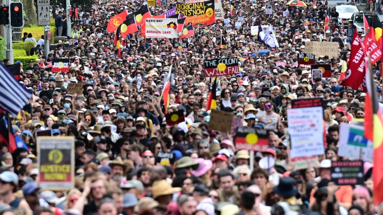 A large crowd of demonstrators carrying flags and signs bearing the colours of the Aboriginal flag.