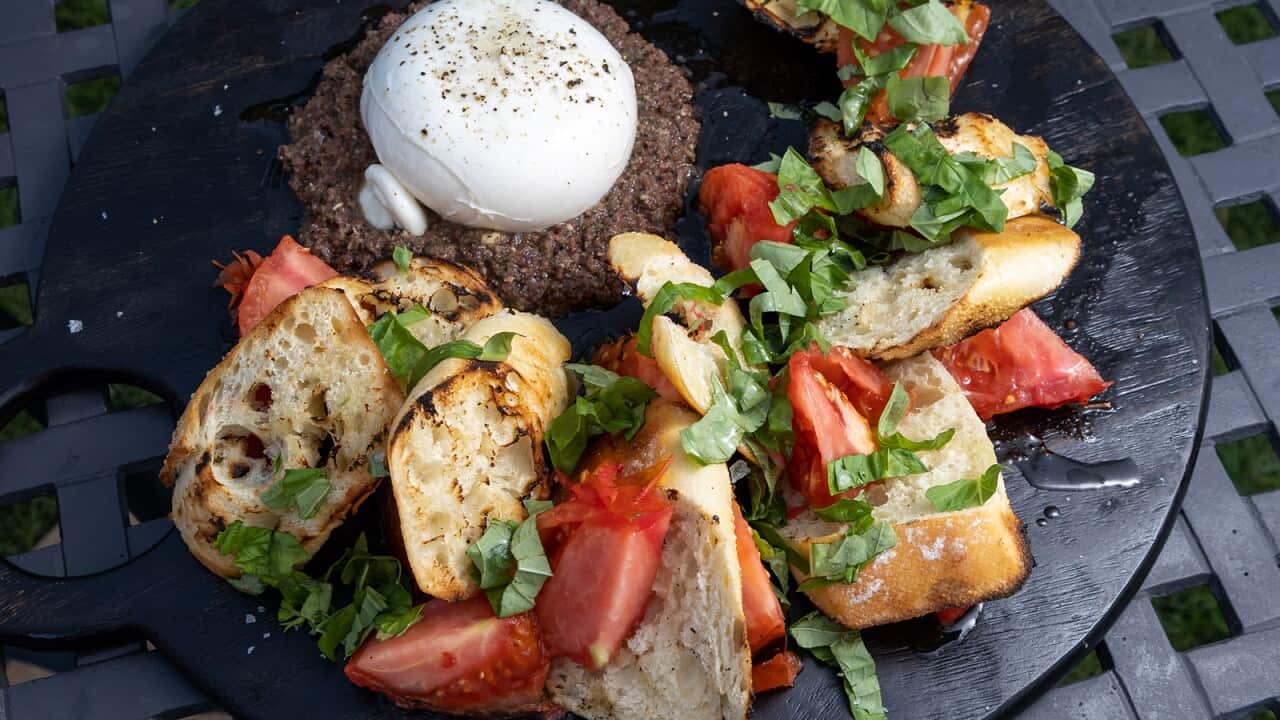 A round blcak tray holds a semiciclue of grilled bread and tomato, garnished with basil, sitting around a white ball of cheese on a round bed of olive dip.