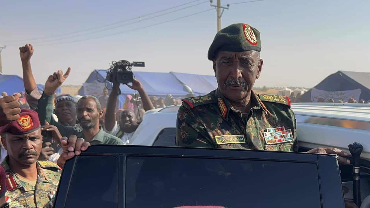An army official is standing in an SUV with the door ajar. He's surrounded by army personnel, and a cameraperson is filming the scene.