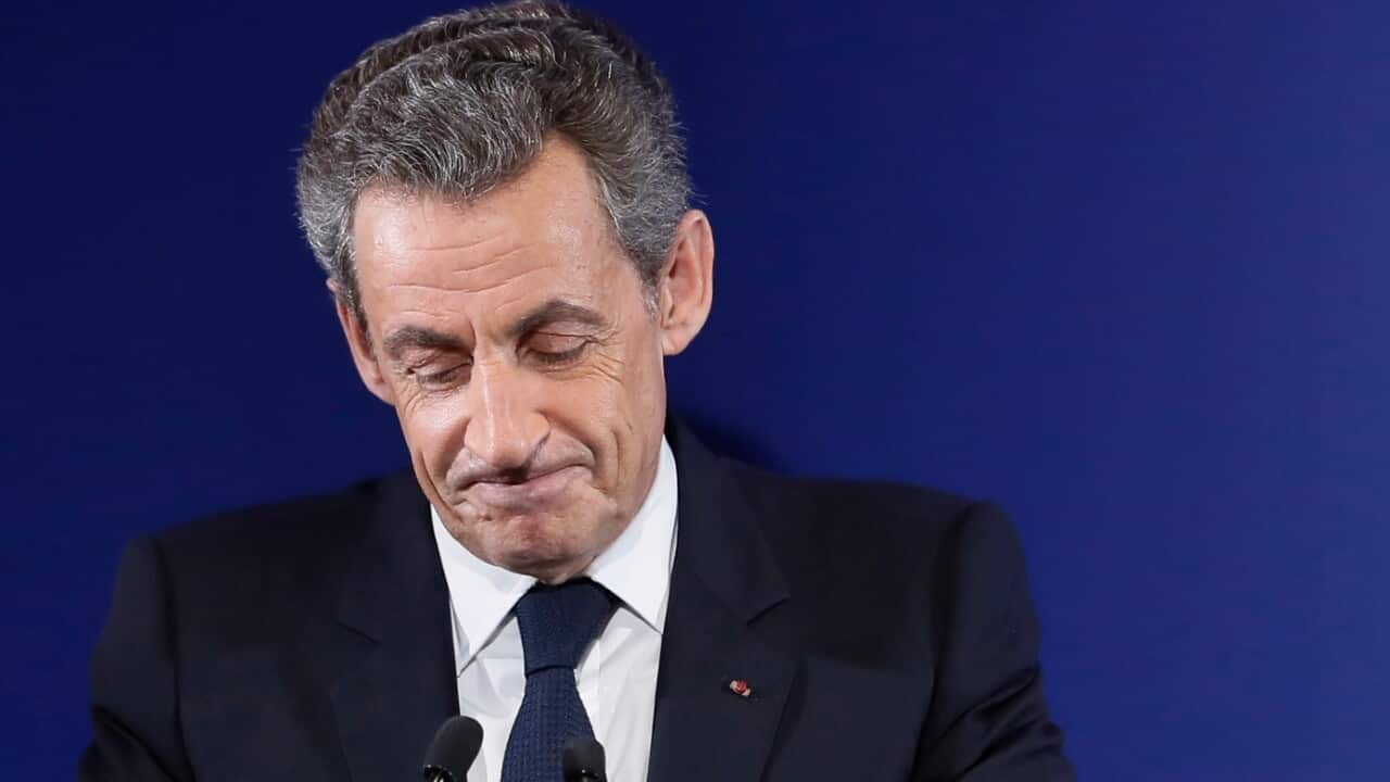 A middle-aged man wearing a suit frowns before a microphone. 