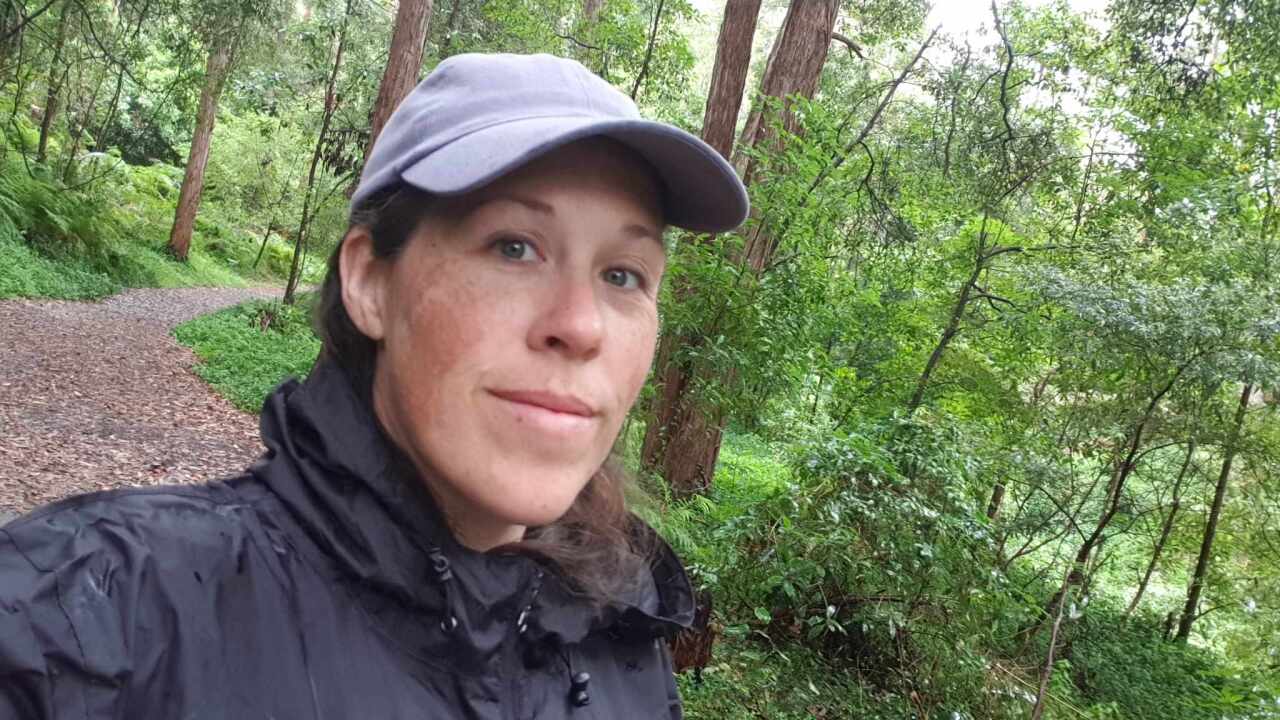A woman in a forest wearing a black jacket and grey cap