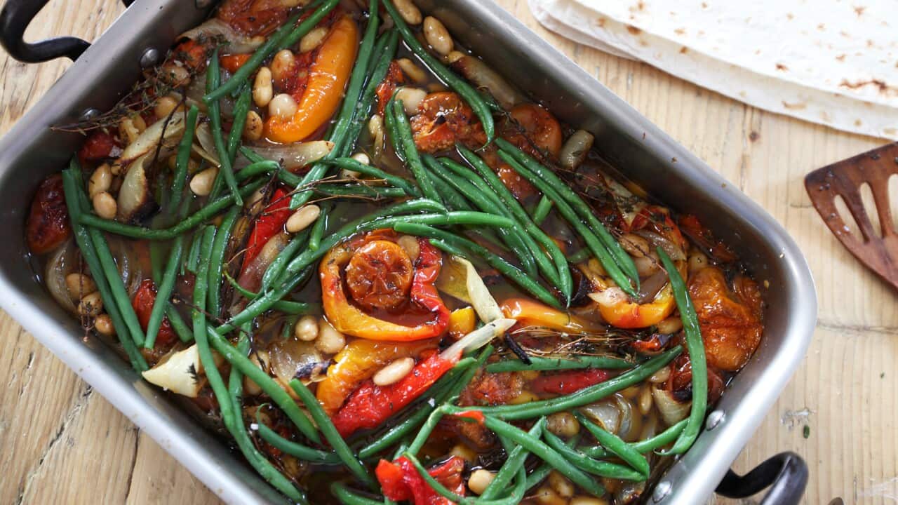 A silver baking tray with black handles holds a bright vegetable stew, with long green beans and red capsicum pieces adding to the colour. The edge of a stack of flatbreads can be seen alongside.