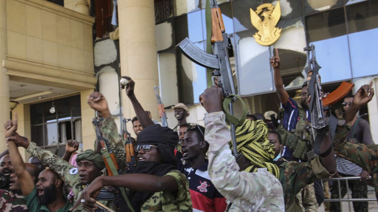 A group of armed men in military and paramilitary clothing raise their weapons and fists in the air in front of a damaged building with a gold emblem featuring a bird. The building's windows are shattered, and its facade shows signs of destruction.