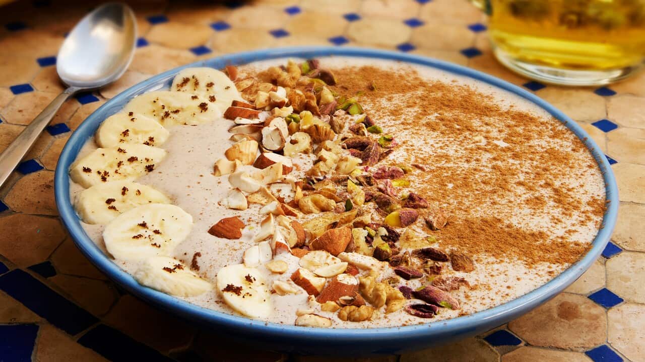 A blue rimmed bowl sits on a tiled surface. It has a smoothie mixture in it, topped with a row of banana slices, nuts and cinnamon.