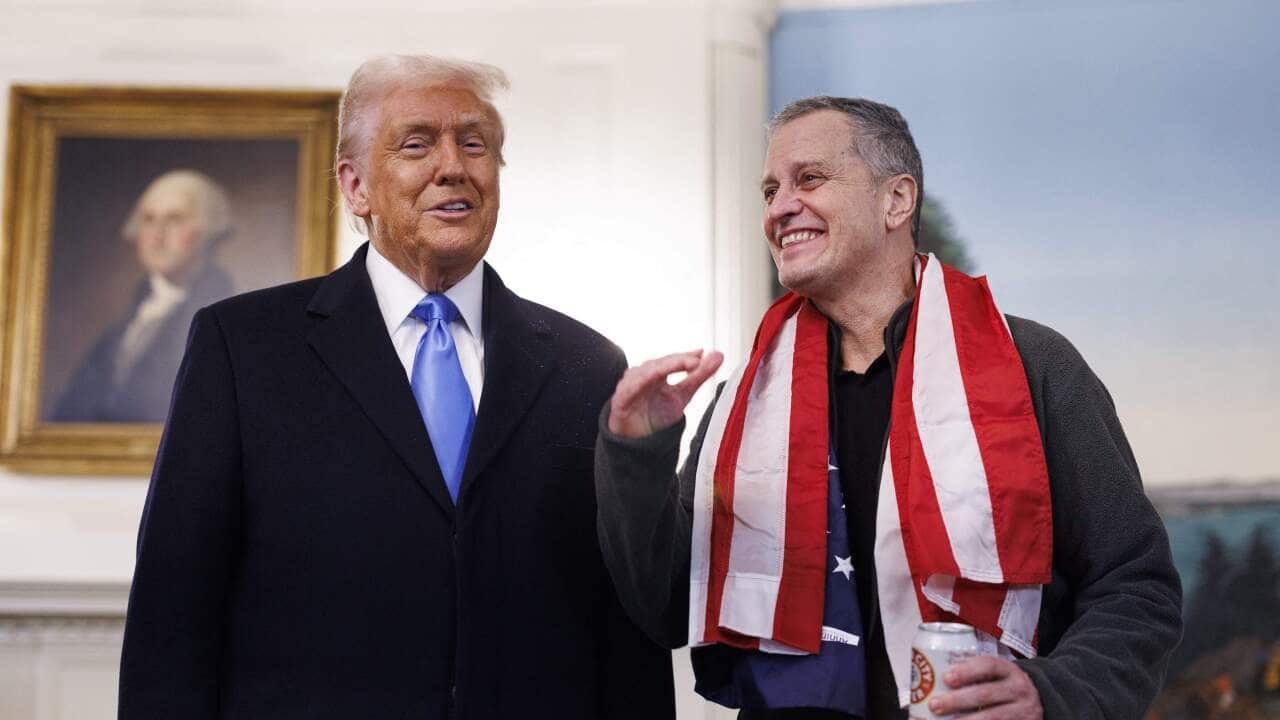 A man in a suit and tie on the left smiles as he talks to another man who has the American flagged draped around his shoulders
