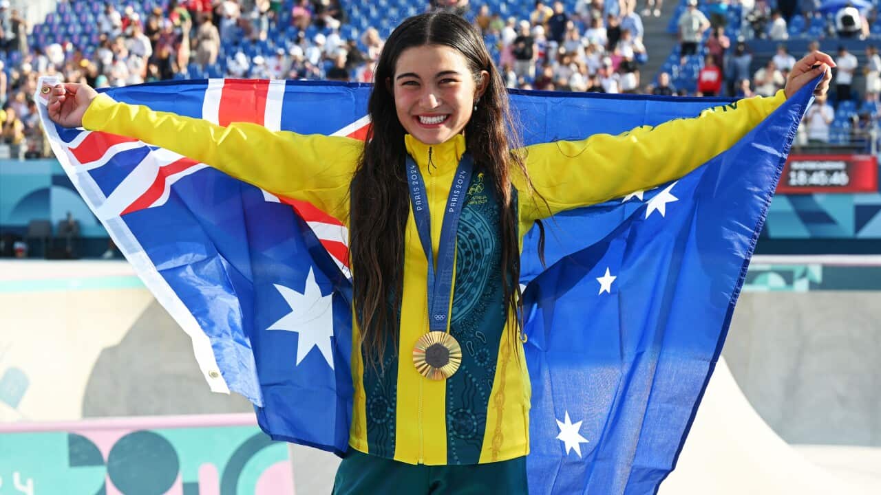 A girl wearing a green and gold tracksuit and with a gold medal around her neck holds up an Australian flag behind her