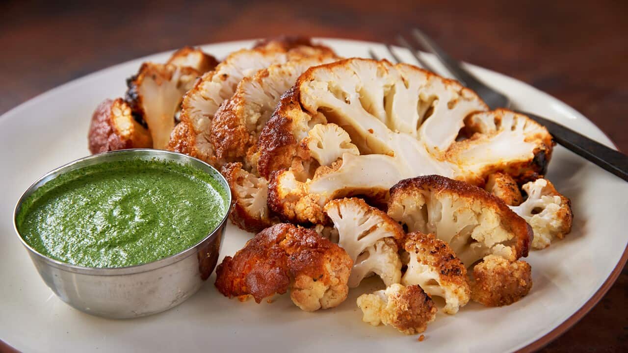 Slices of cauliflower with reddish edges sit on a wide platter, along with a metal bowl filled with a bright green dip.