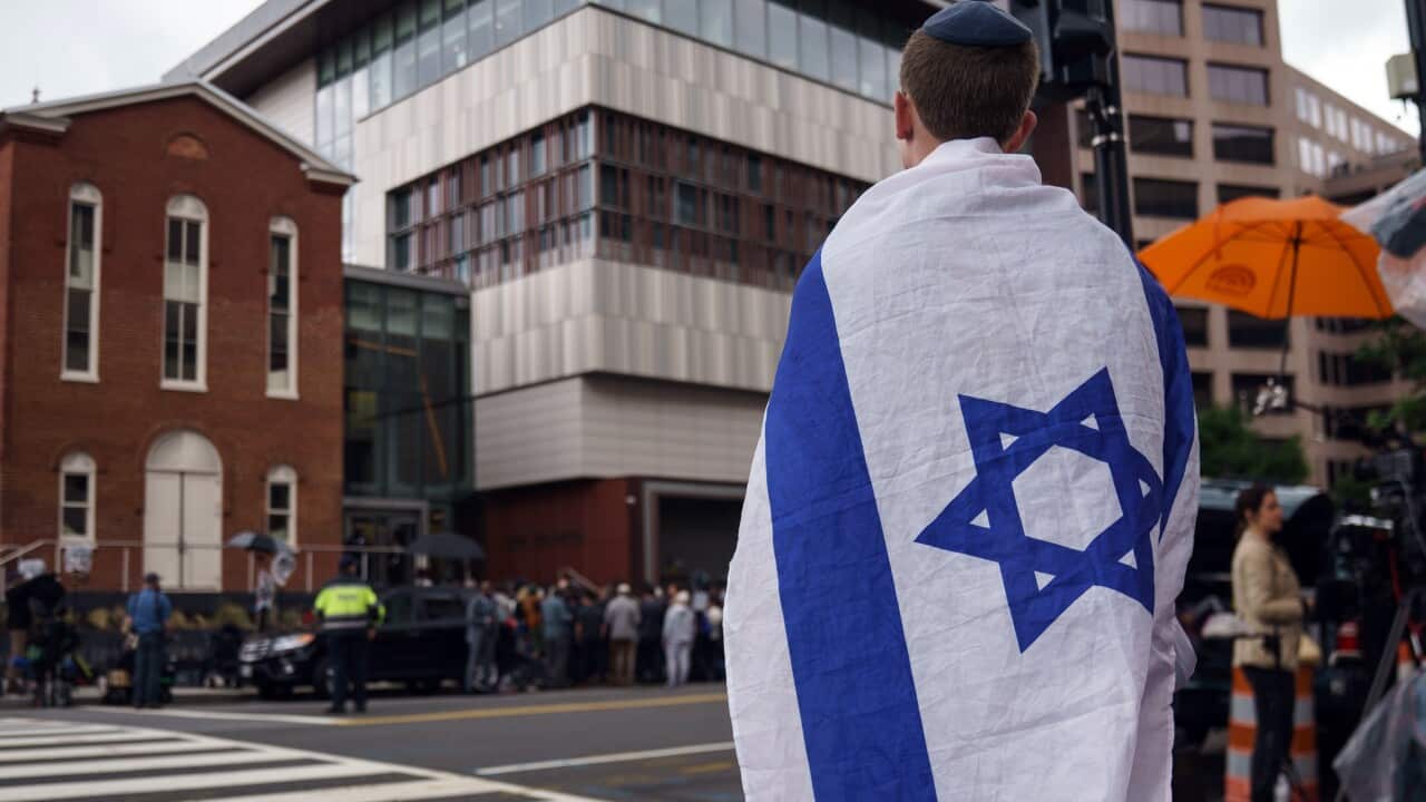 A man with the Israeli flag draped over his shoulders looks at a crime scene across the road, where a crowd is standing.