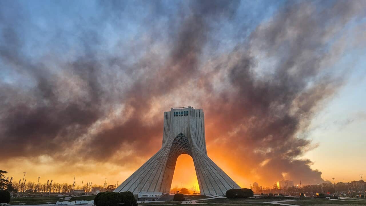 The Azadi Tower in Tehran stands silhouetted against a dramatic sunset, with thick smoke billowing behind the iconic arched monument.