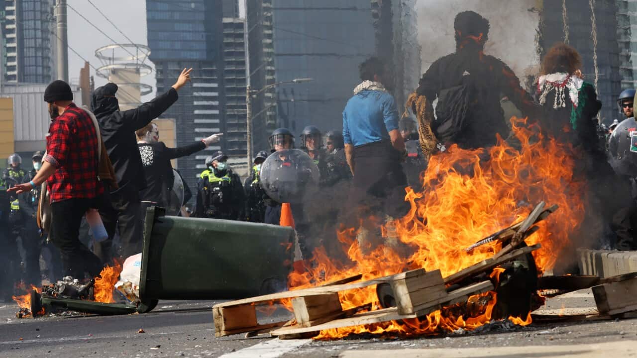 A pallet and bin shown on fire as protesters clash against police in the middle of a road.