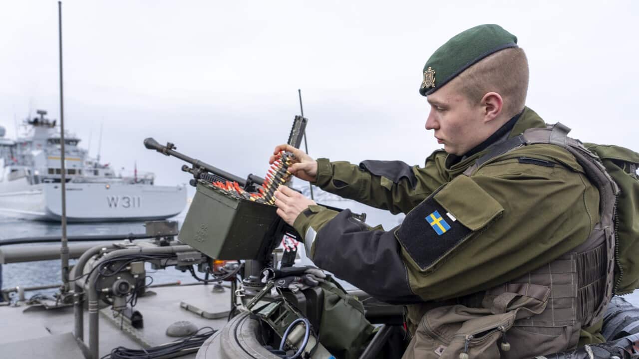 A soldier in green uniform and green beret loads bullets into a gun magazine from a turret on a boat. He has a small Swedish flag badge sown onto the left upper arm of his uniform