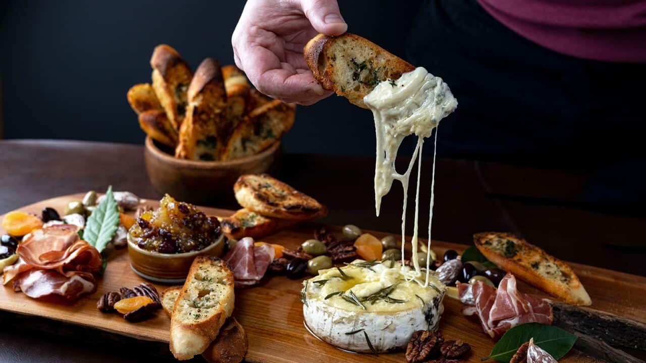 A hand holds a thin piece of toasted bread, with cheese melting off it, above a long wooden platter which holds a baked round of cheese and other items, including olives, cured meat and pecans.