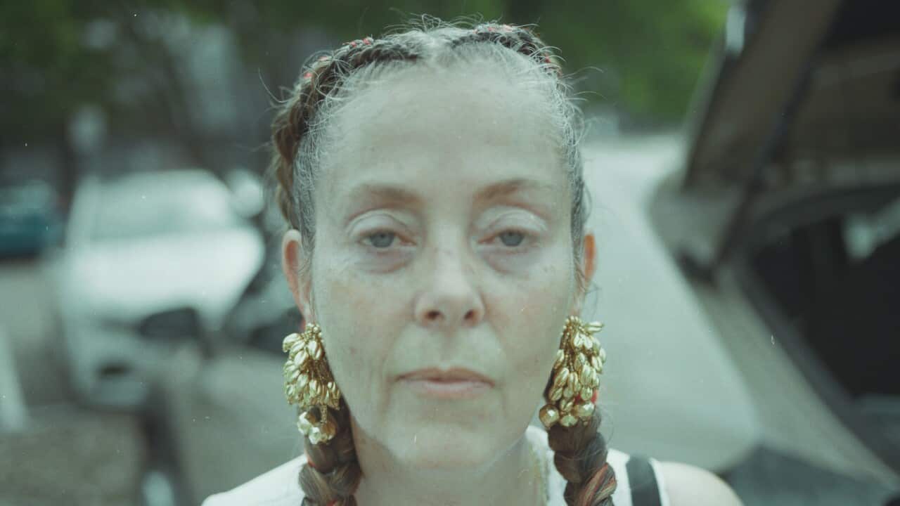 a film photograph of a woman standing in a carpark in a crotchet white dress with her hair in braids with red streaks and gold earrings