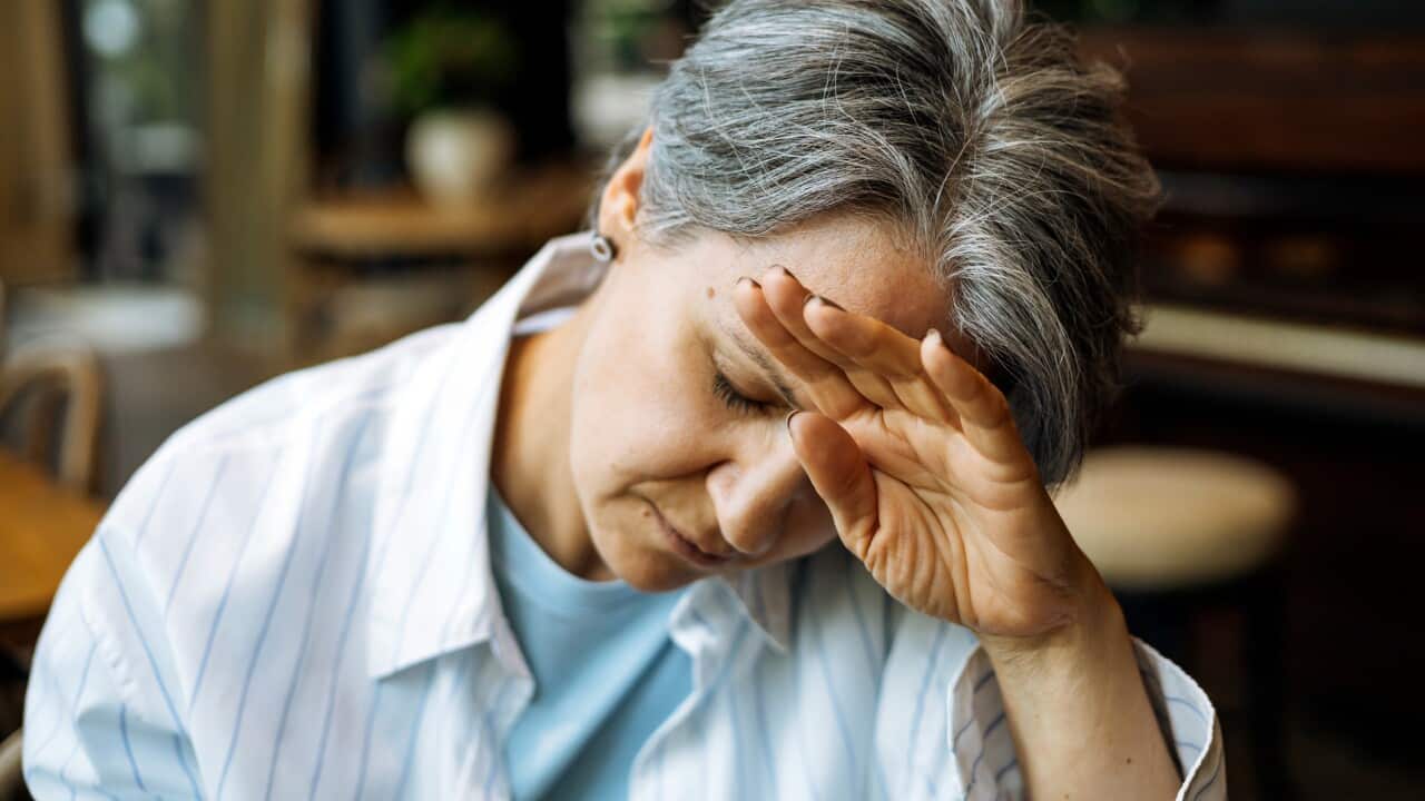 An older woman with gray hair, covering her face with her hand, seems to be experiencing a moment of stress.