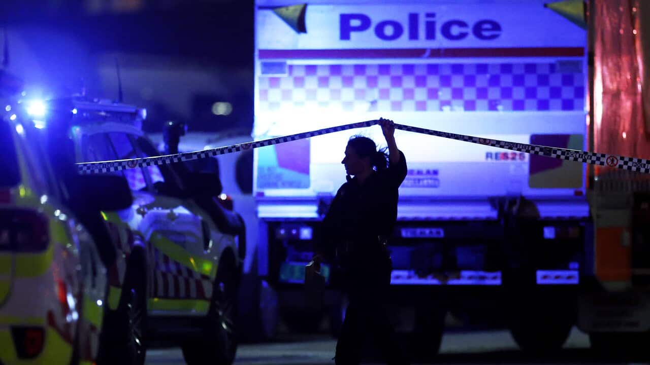 A police officer lifts crime-scene tape while walking between emergency vehicles illuminated by blue lights at night.