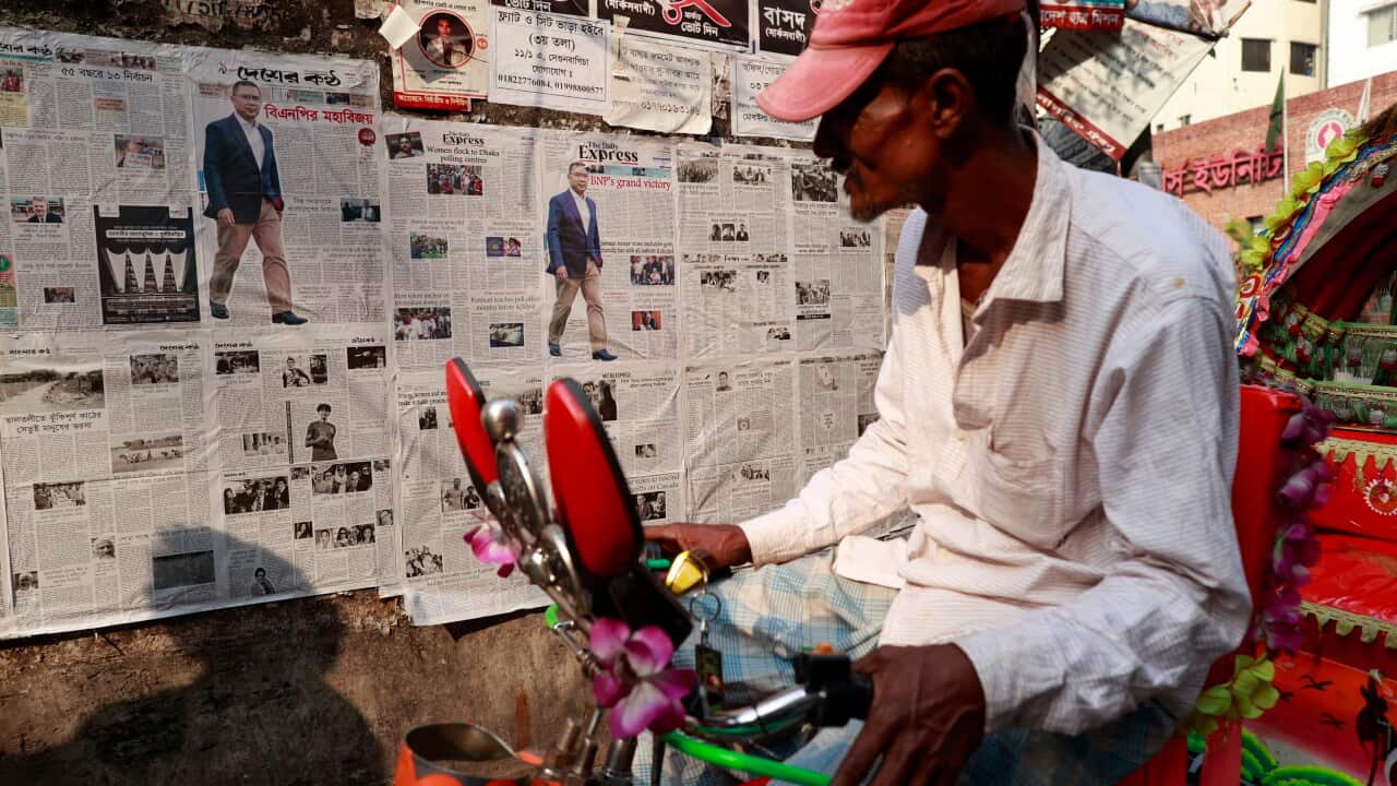 A man on a bike reads a newspaper pasted on a roadside wall.