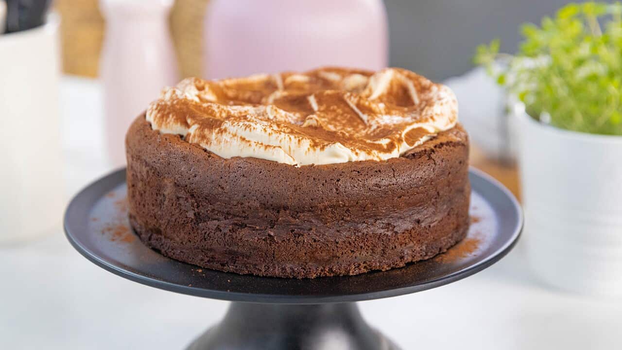 A deep chocolate cake, with a creamy topping, sits on a black cake stand.