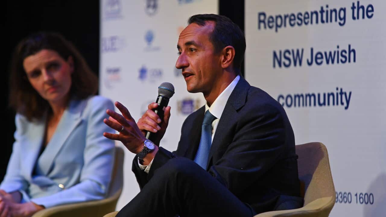 A man wearing a dark suit is sitting on a stage, speaking into a microphone he is holding next to a woman in an aqua-blue blazer.