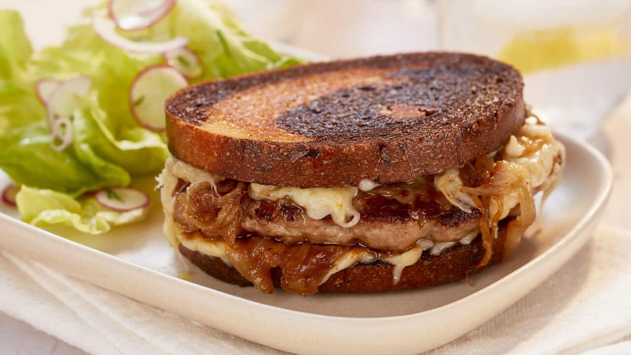 A toasted sandwich, filled with a meat patty, cheese and caramelised onion, sits on a white rectangular plate, which sits on a white napkin. Green lettuce leaves sit behind the sandwich.