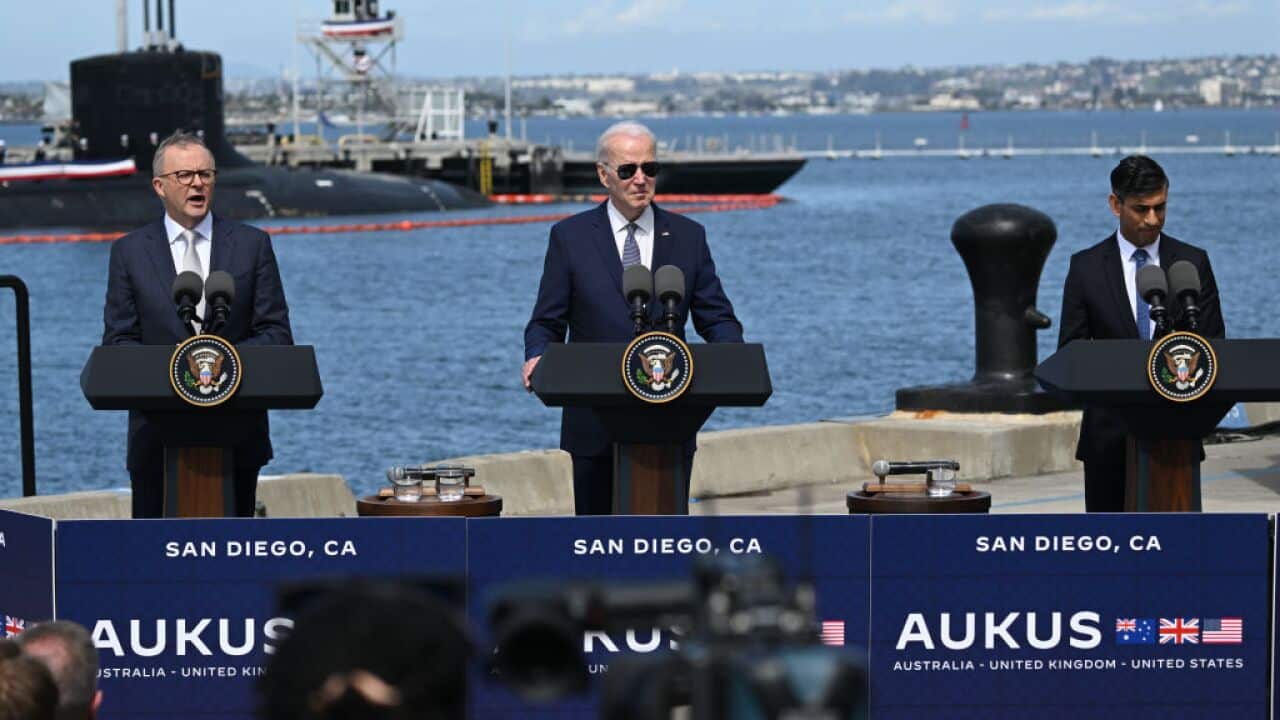Three men standing at lecterns on a dock. There is a submarine in the water behind them