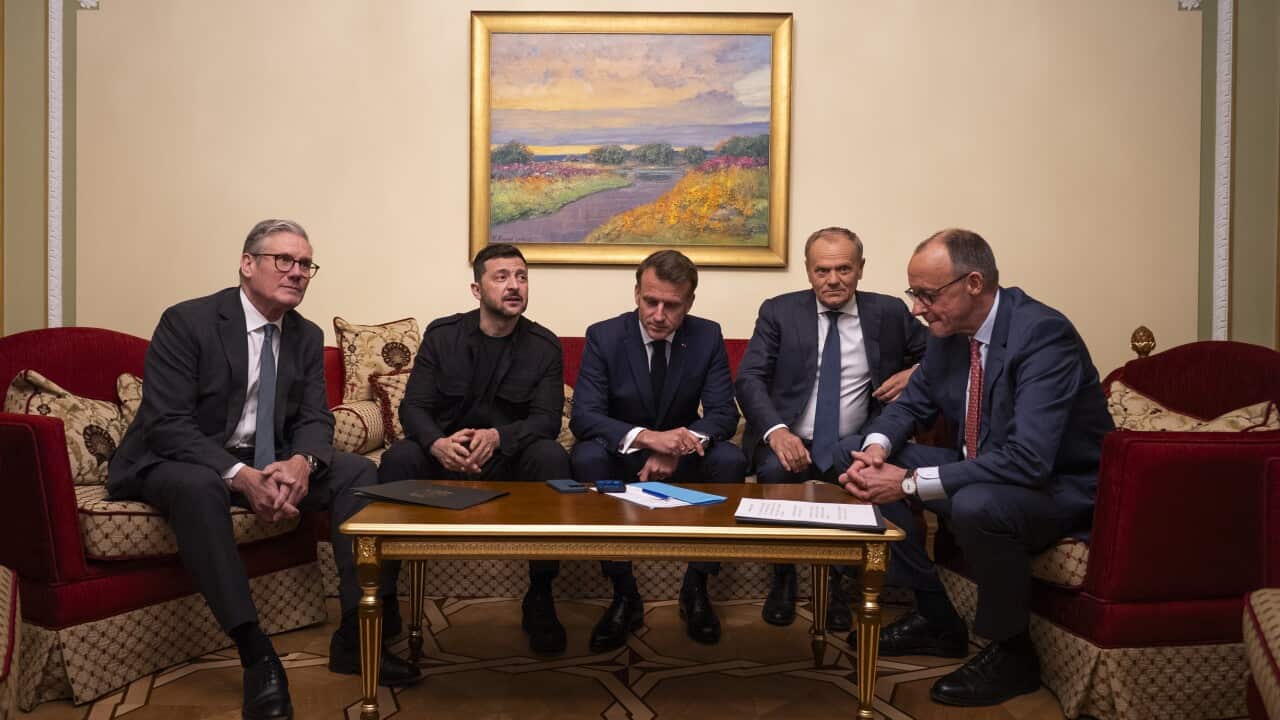 Five men in suits seated on chairs. A phone on the desk in front of them is on speakerphone, and they are listening.