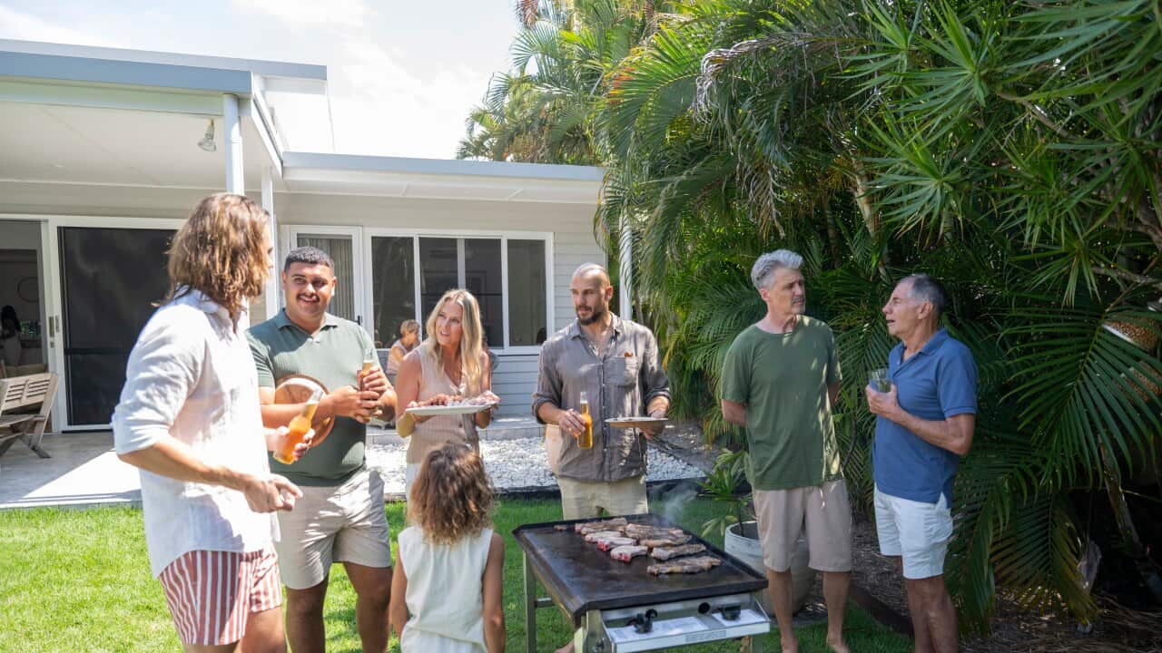 Diverse group of men, women, and children standing in sunny back yard of modern one-story home, talking and drinking as meat cooks on grill.