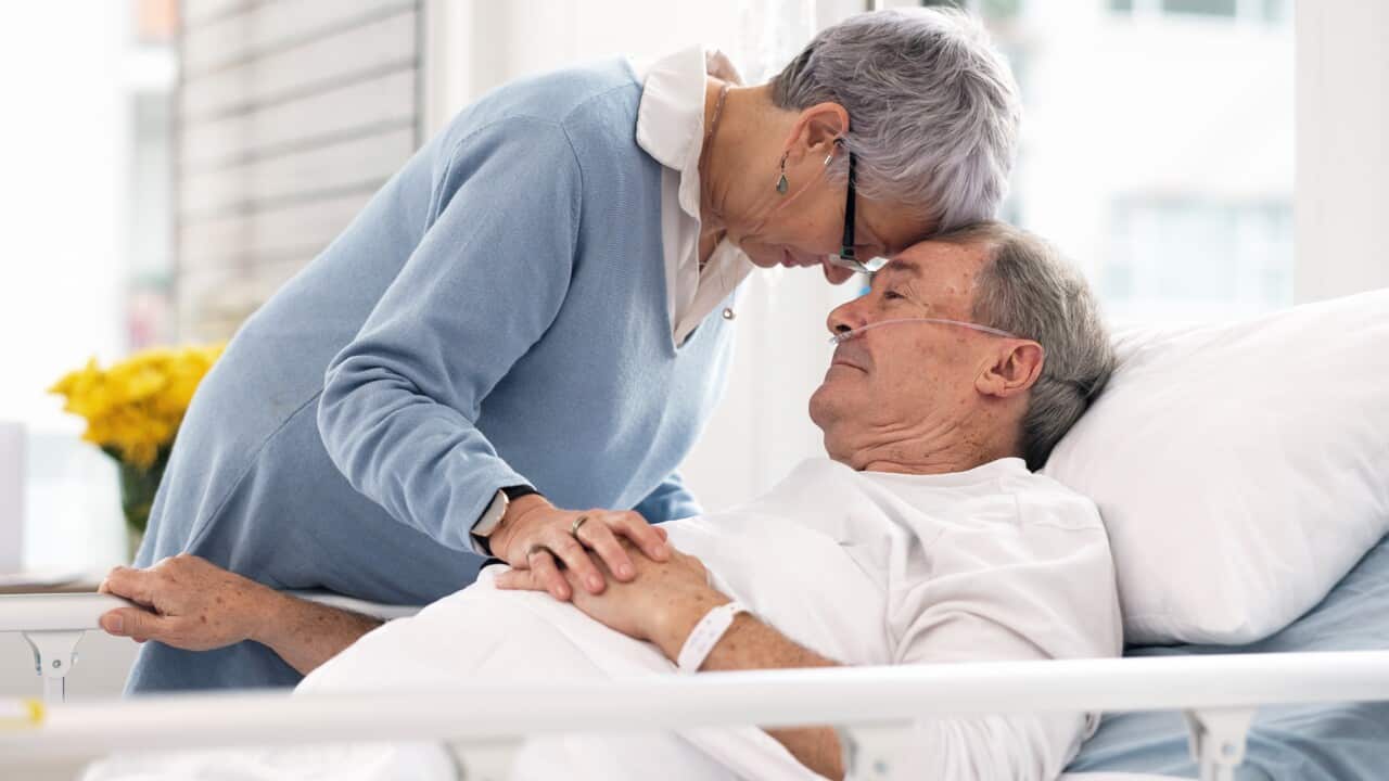 An elderly woman in a light blue blouse rests her head against that of an elderly man lying in a hospital bed with tubes running into his nose.