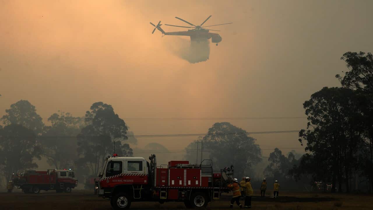 Firetrucks and a helicopter dumping water are visible through thick bushfire smoke.
