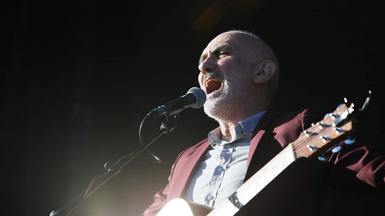 A man in a red blazer and blue shirt playing a guitar and singing into a microphone