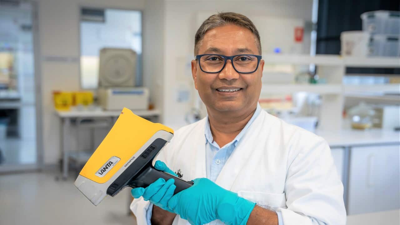 A man in a white lab coat holds a yellow handheld scanner in a science lab.