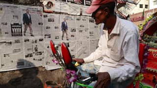 A man on a bike reads a newspaper pasted on a roadside wall.