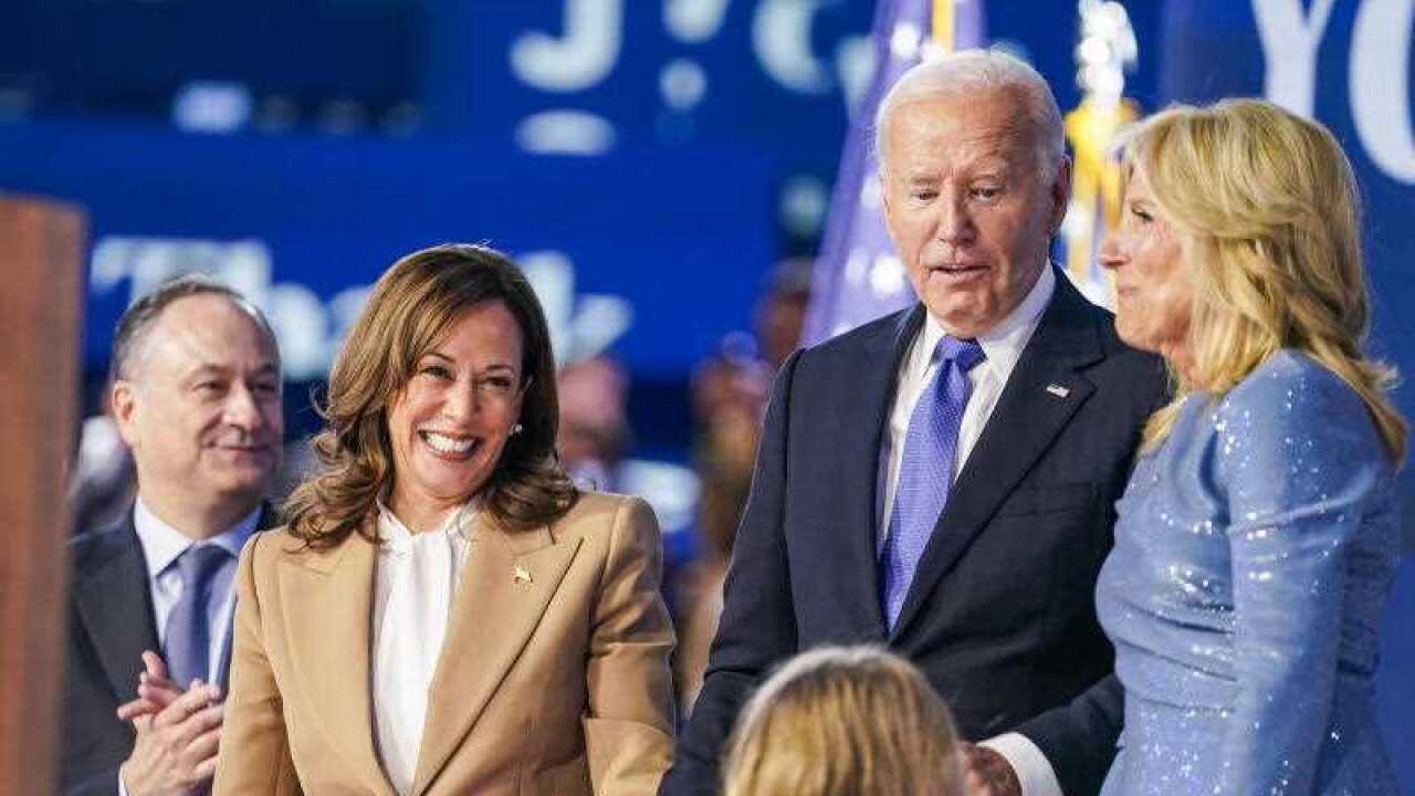Kamala Harris, wearing a tan suit, and Joe Biden, wearing a dark suit and blue tie and standing next to his wife, who is wearing a blue dress. There is a child facing them.