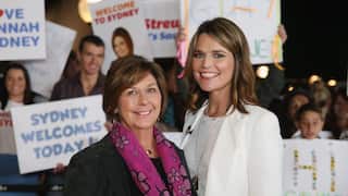 Two woman smiling side by side. In the background, people can be seen carrying signs reading "Welcome to Sydney" and "Sydney welcomes Today!"