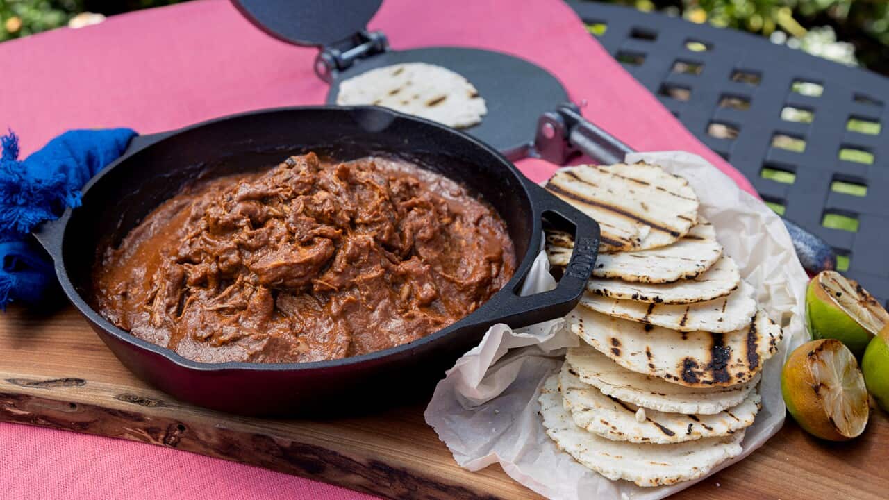 A cast iron pot holds a rich brown chicken mixture. Tortillas with grill marks sit alongside.