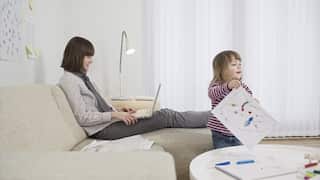 A white woman using a laptop on her lap as she sits on a couch, her young daughter nearby is drawing on paper.