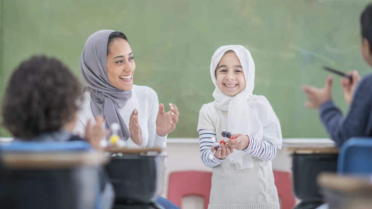 Teacher wearing hijab clapping young girl who is doing a presentation in class.