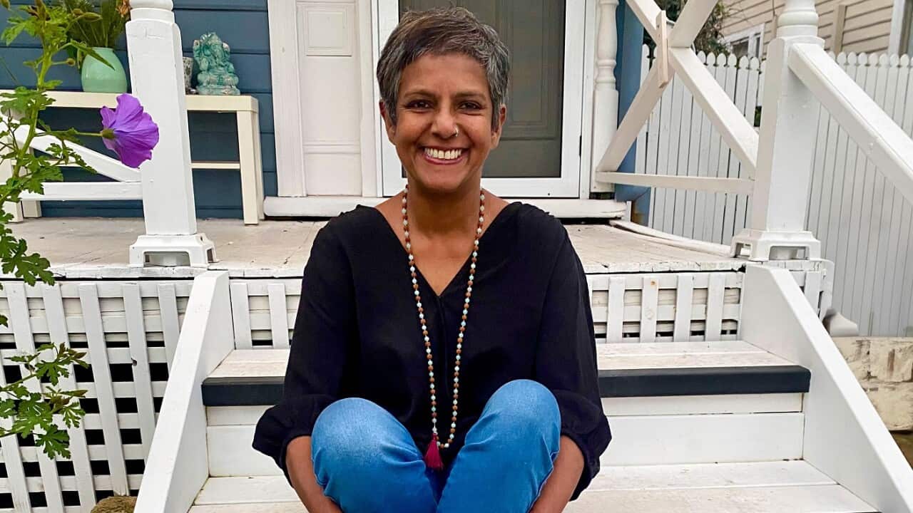 A smiling person with short grey hair, wearing a black top and blue pants, is sitting on the steps of a white wooden porch.