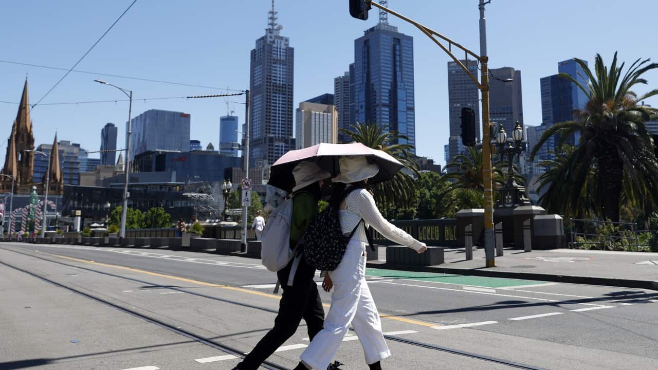 Two people wearing hats, backpacks, and long sleeve pants and shirts walk across the street while holding a sun umbrella, crossing the street. City buildings can be seen behind them.