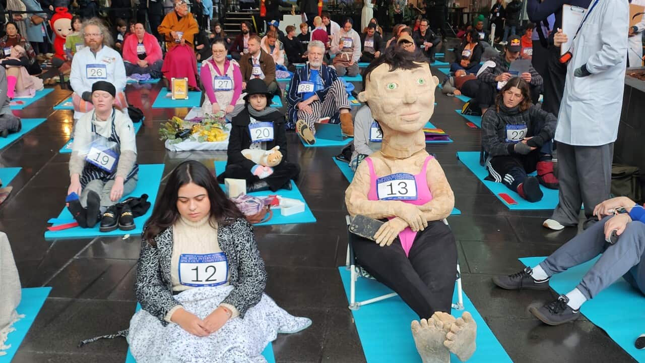 Rows of people sitting on blue yoga mats in costumes. In the foreground one person is wrapped in a blanket and another wears a large paper mache suit.
