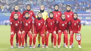 A team of female soccer players from the Iranian national team, wearing red kits and black hijabs, poses for a group photo on a rainy pitch before a match.