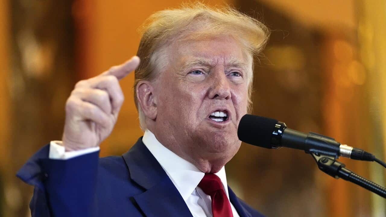 Donald Trump wearing a blue suit, white shirt and red tie, speaking animatedly in front of a microphone.