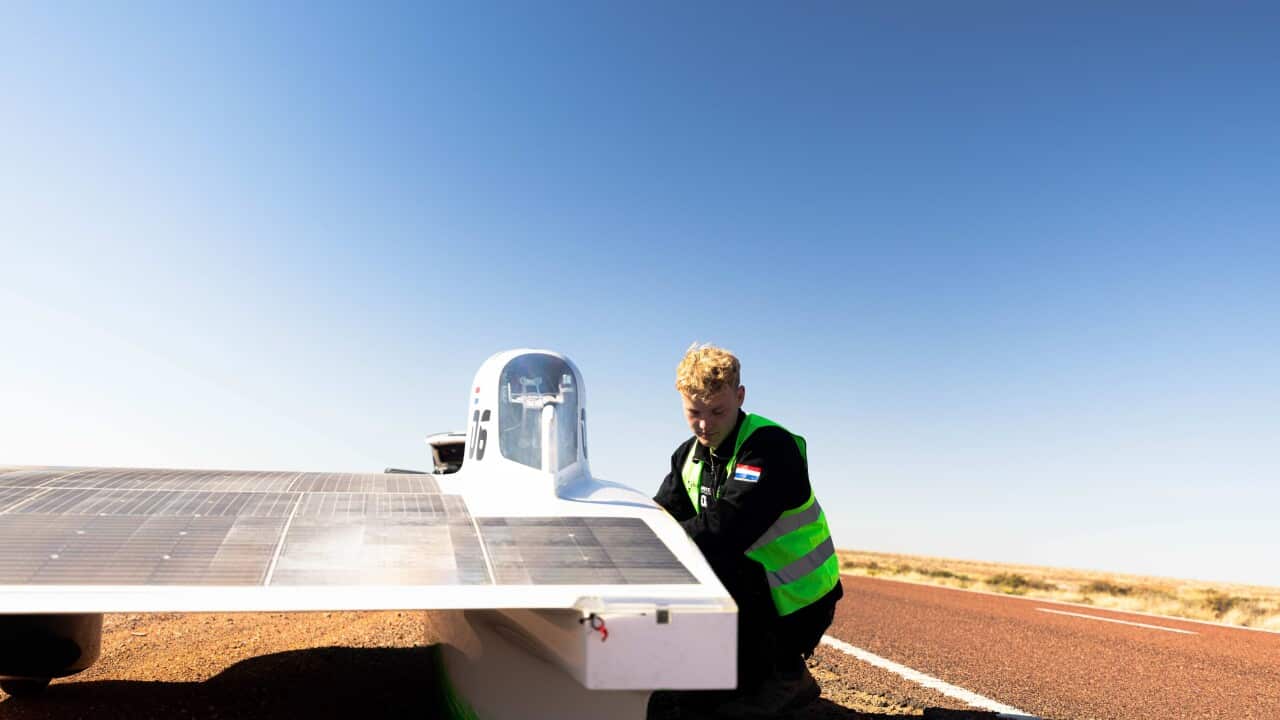 A person wearing a hi-vis vest over his clothes, squatting next to a solar vehicle parked on the side of an outback road.