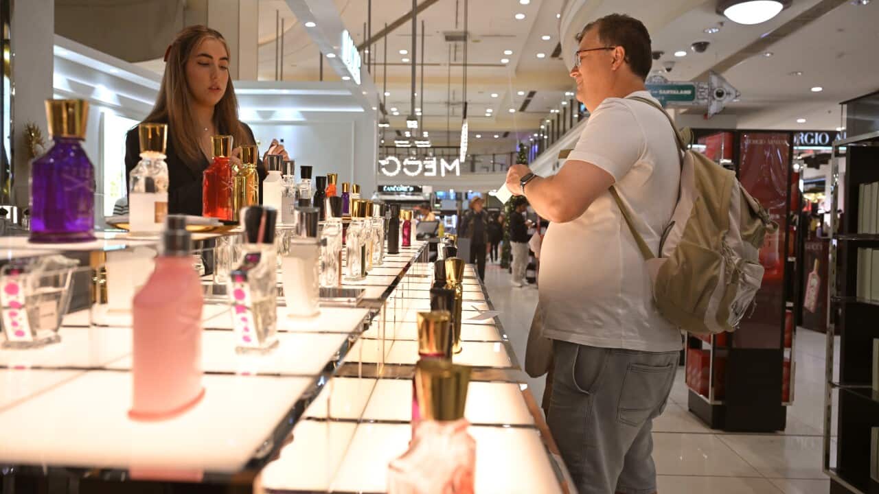 A man in a white T-shirt and grey shorts, carrying a backpack, stands next to a counter lined with rows of perfume bottles, while a saleswoman stands behind it.