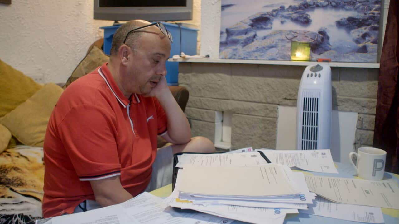 A man in an orange polo shirt stares at a table laden with papers and a boy sitting by his side