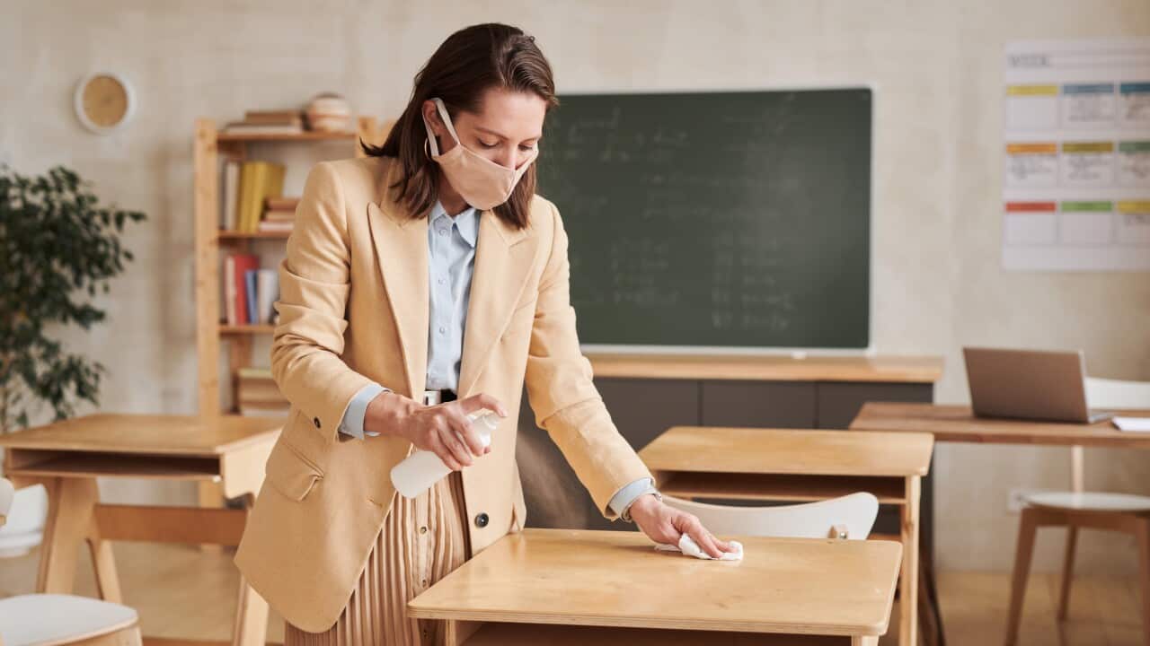 Teacher wearing mask and sanitising desks in school classroom as students prepare to go back to school.
