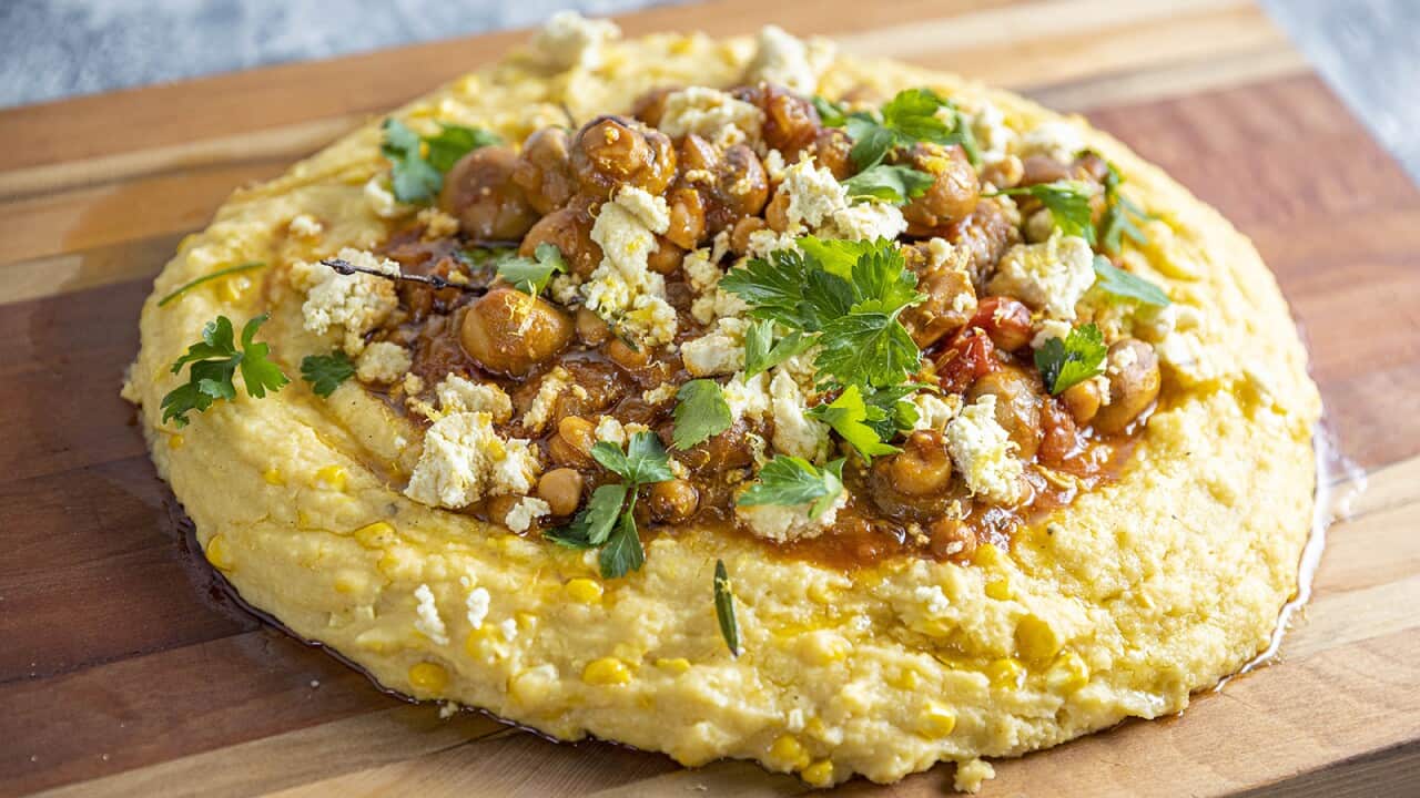 A round mound of polenta sits on a wooden board, topped with a cooked mushroom and chickpea mixture, garnished with parsley leaves.