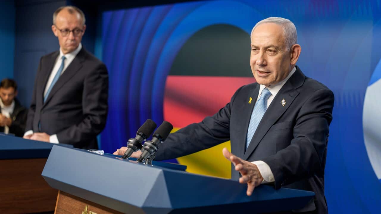 A man in a suit speaks at a podium with another man in a suit looks at him