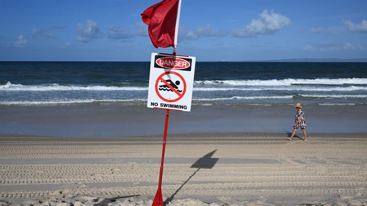 A red flag and a sign reading “danger, no swimming“ on a beach, with waves in the background and a person walking along the shore.