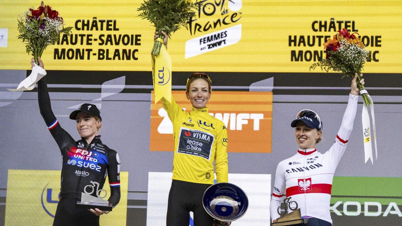 Three women cyclists stand on the podium holding medals in one hand and bouquets in the other hand raised in victory.